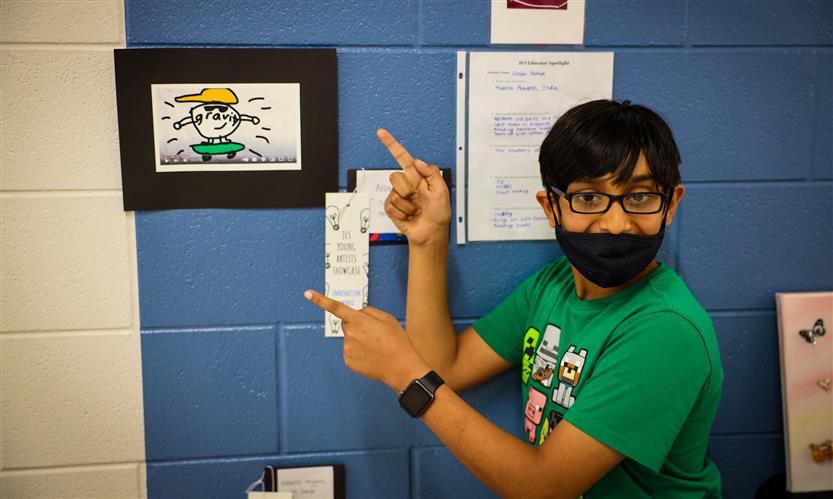 A boy wearing green points at a screenshot hanging on the wall